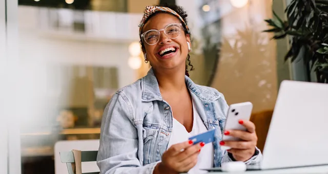 Woman holds a credit card whilst sitting in front of her laptop and is laughing